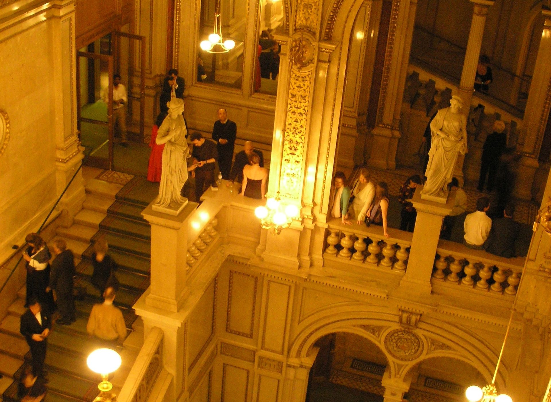 Elegante Menschen in Abendgarderobe gehen und versammeln sich auf kunstvollen Treppen in einer großen, warm beleuchteten Halle mit klassischer Architektur, dekorativen Statuen und komplizierten Details an den Wänden.