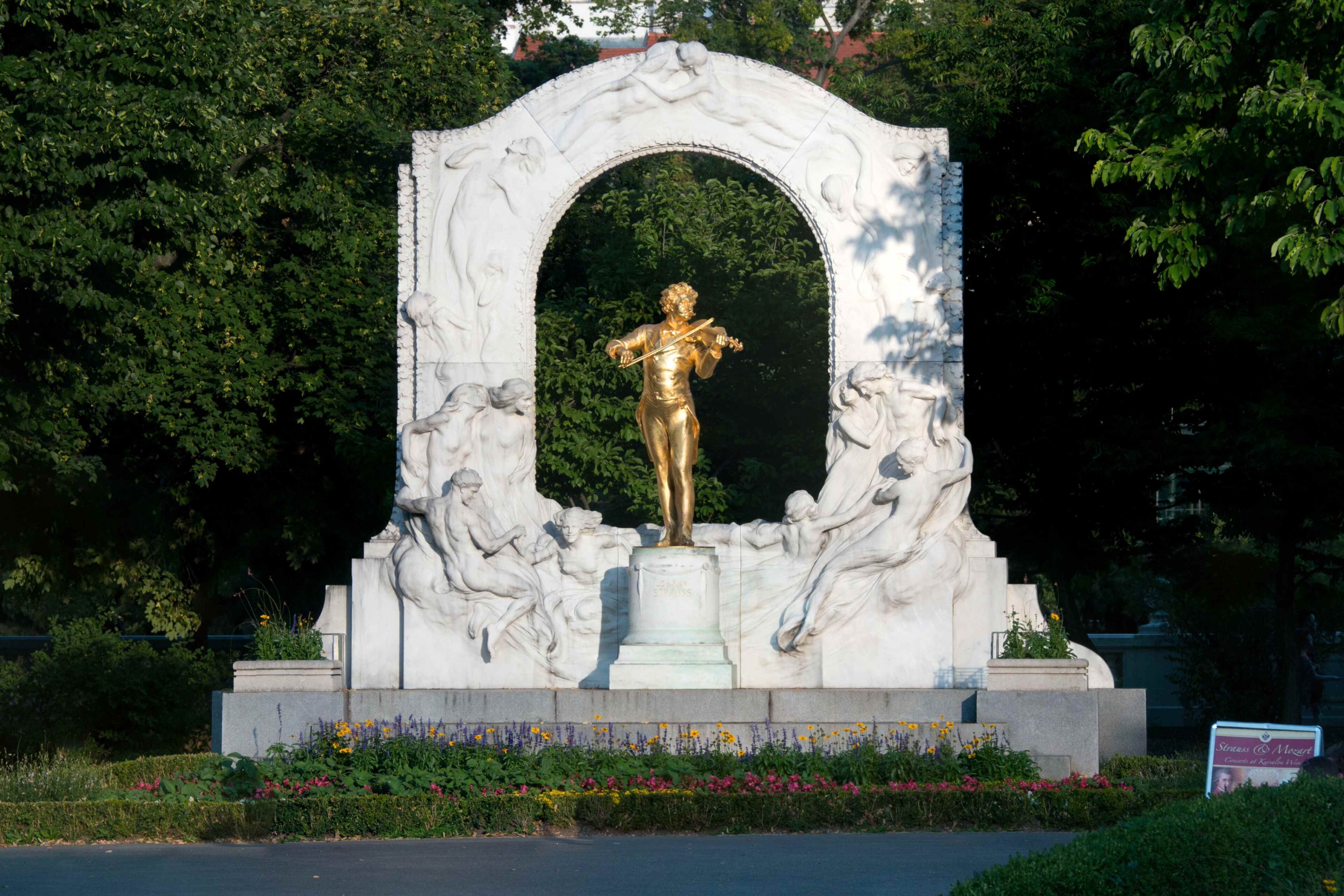 Eine goldene Statue des Geigers Johann Strauss steht unter einem weißen Steinbogen, der mit Skulpturen geschmückt ist, umgeben von Grün und bunten Blumen in einer Parklandschaft.