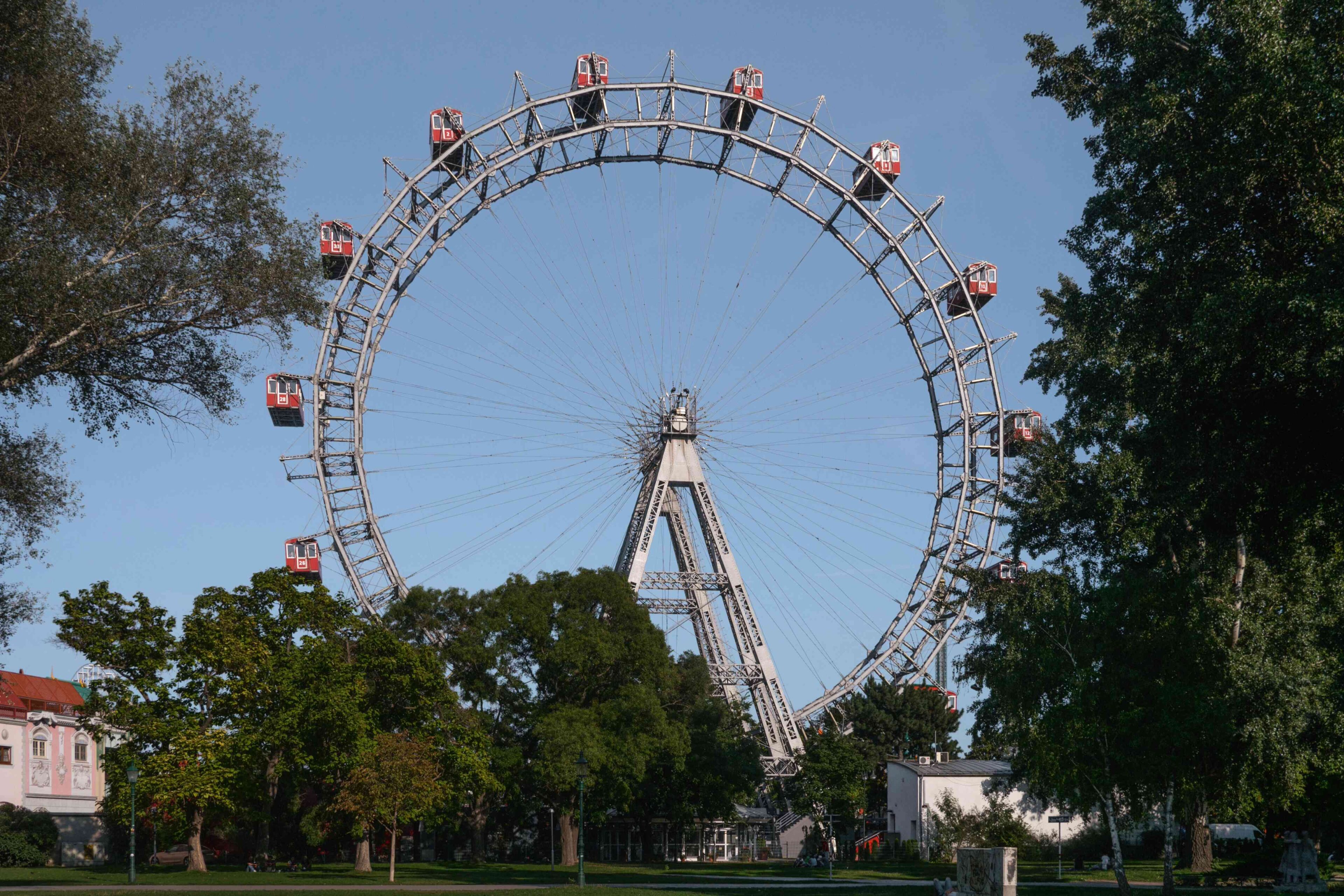 Ein großes Riesenrad mit roten Kabinen steht zwischen Bäumen und Grün unter einem klaren blauen Himmel.