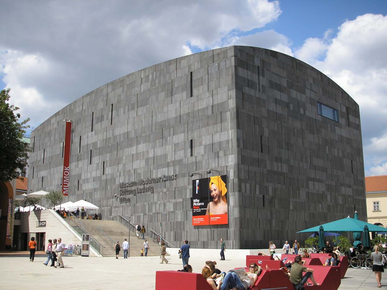 Ein großes, modernes graues Steingebäude mit minimalen Fenstern, das als Museum für zeitgenössische Kunst Mumok bezeichnet wird. Menschen sitzen auf roten Bänken auf einem sonnigen Platz mit Regenschirmen und einer Werbetafel davor.