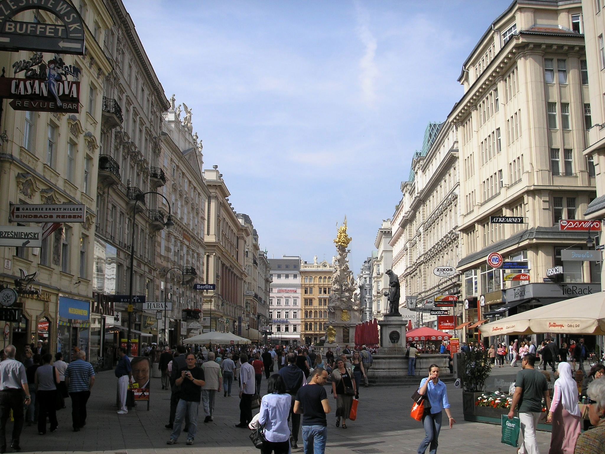 Eine belebte Fußgängerzone mit historischen Gebäuden, Geschäften und Straßencafés in Wien, Österreich, mit spazierenden Menschen und einem goldenen Säulendenkmal im Hintergrund.