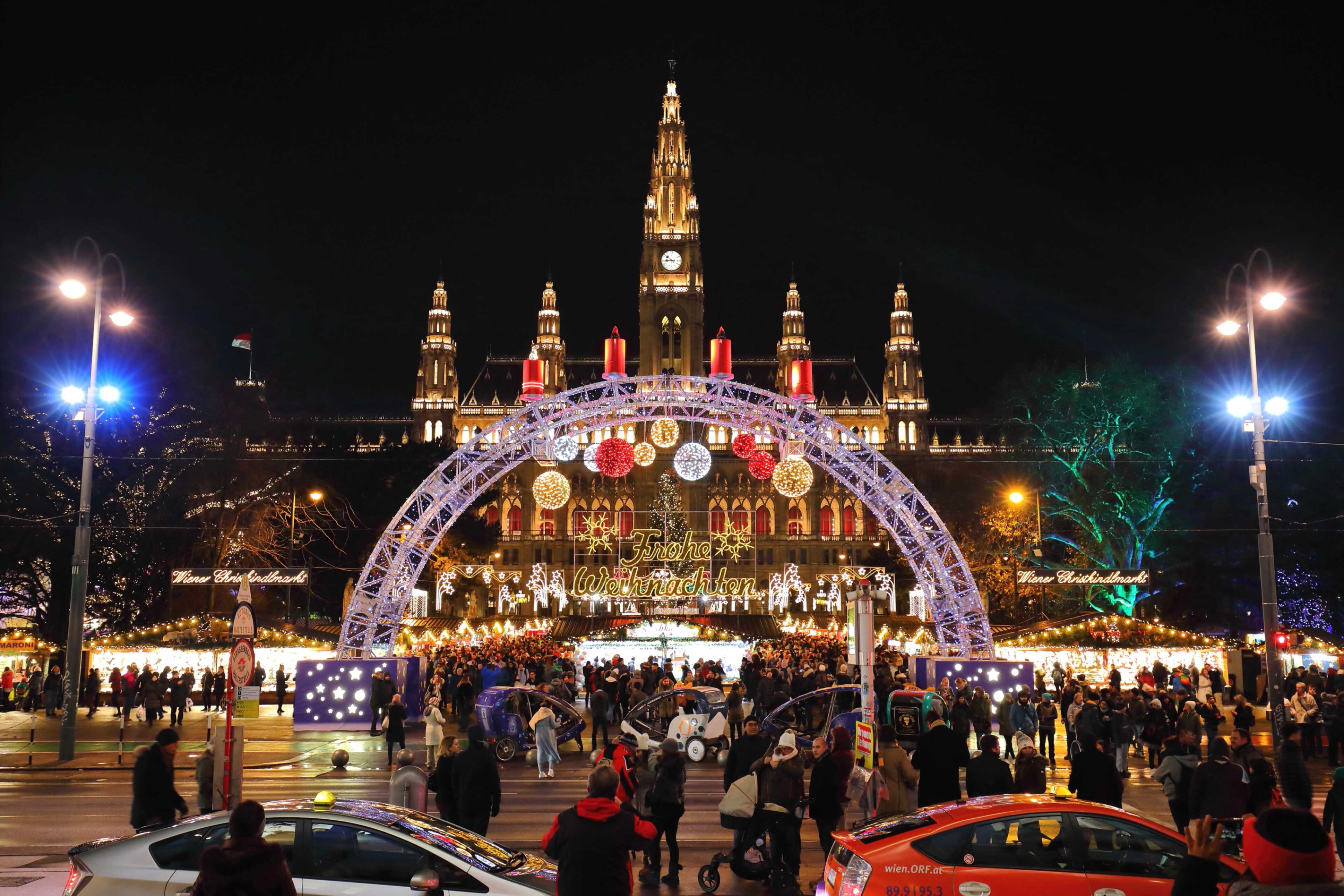 Vor einem großen, beleuchteten historischen Gebäude herrscht nachts ein geschäftiges Treiben auf einem festlichen Weihnachtsmarkt. Im Vordergrund sind bunte Lichter, Weihnachtsdekorationen und vorbeifahrende Autos zu sehen.