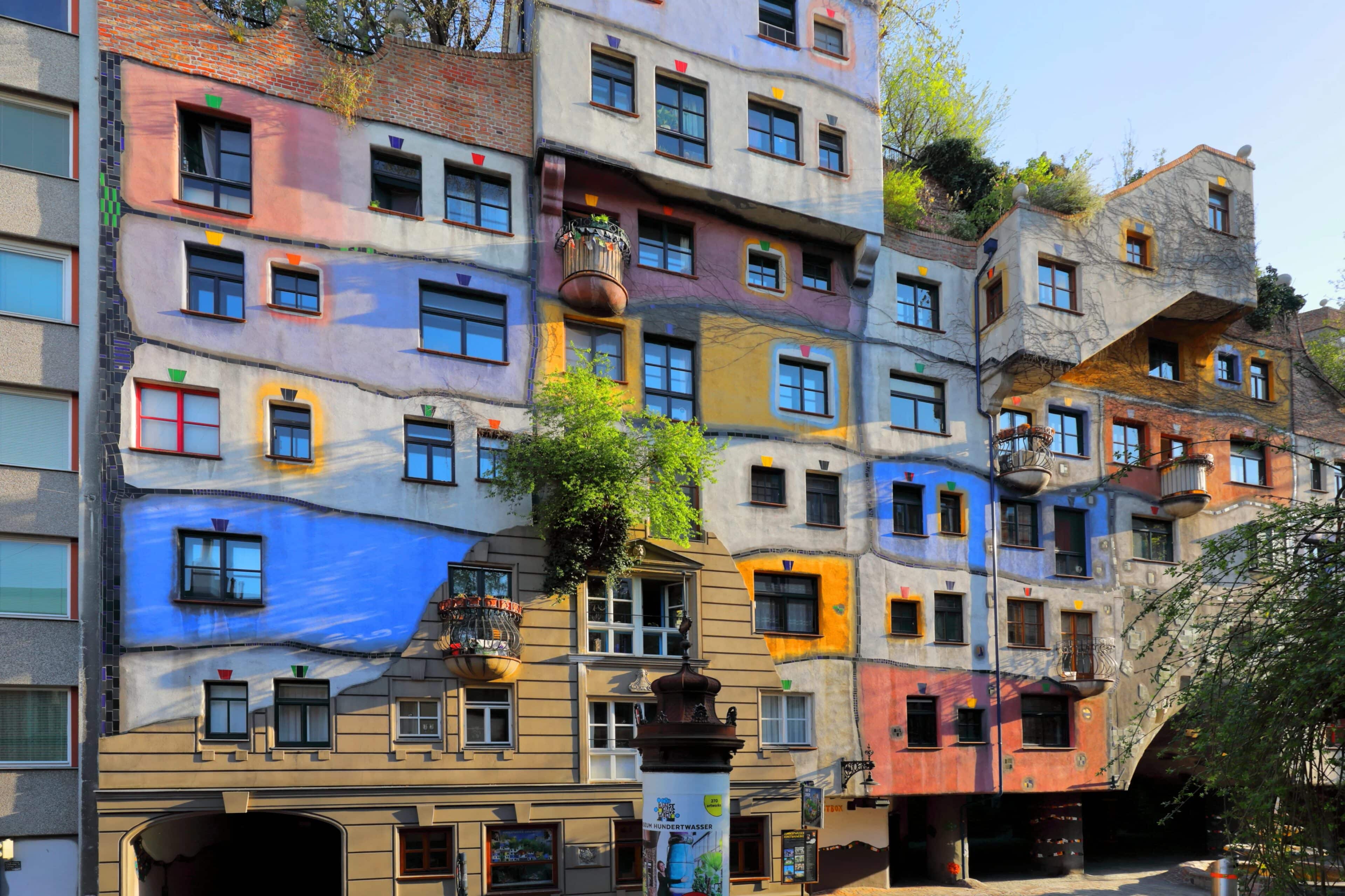 Un colorato edificio residenziale a più piani con finestre di forma irregolare e macchie luminose di rosso, blu, giallo e arancione. La facciata è caratterizzata da verde, elementi artistici e angoli architettonici unici. 