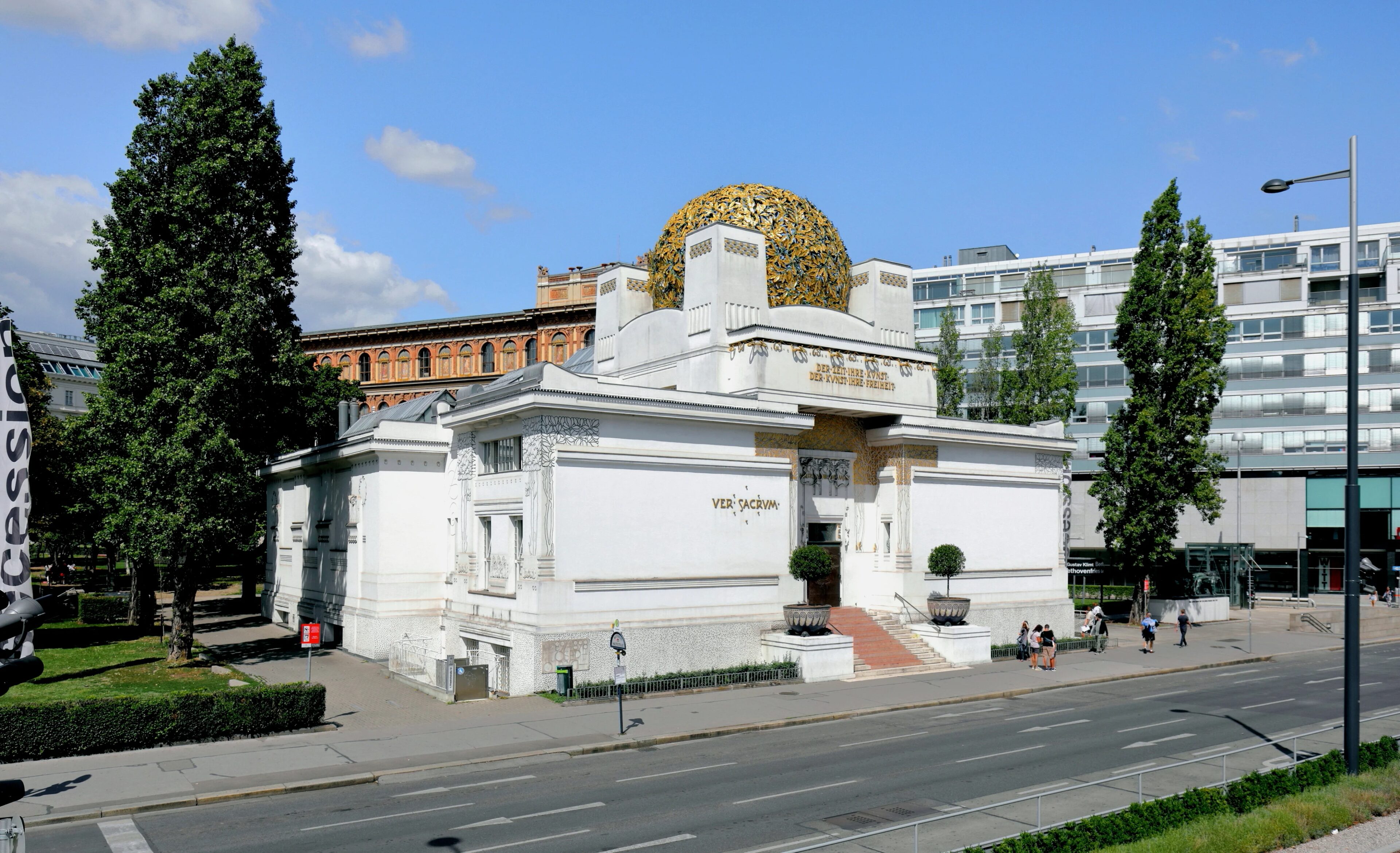 Un edificio bianco con una cupola dorata, noto come l'edificio della Secessione, circondato da alberi ed edifici moderni. Ha una piccola scala che conduce all'ingresso e la scritta "Ver Sacrum" sopra la porta. Alcune persone stanno camminando nelle vicinanze.  