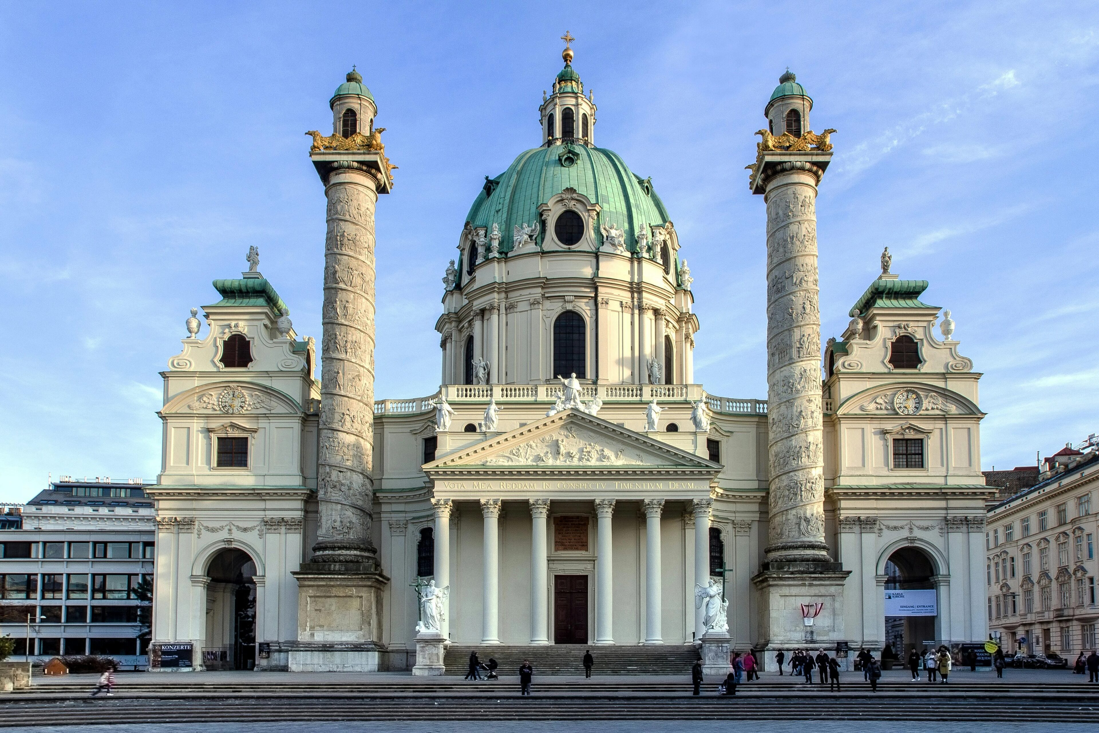 Una grande chiesa barocca con due colonne ornate e una cupola verde si erge sotto un cielo azzurro. La facciata presenta un'architettura classica con colonne e statue. Le persone attraversano la piazza di fronte all'edificio.  