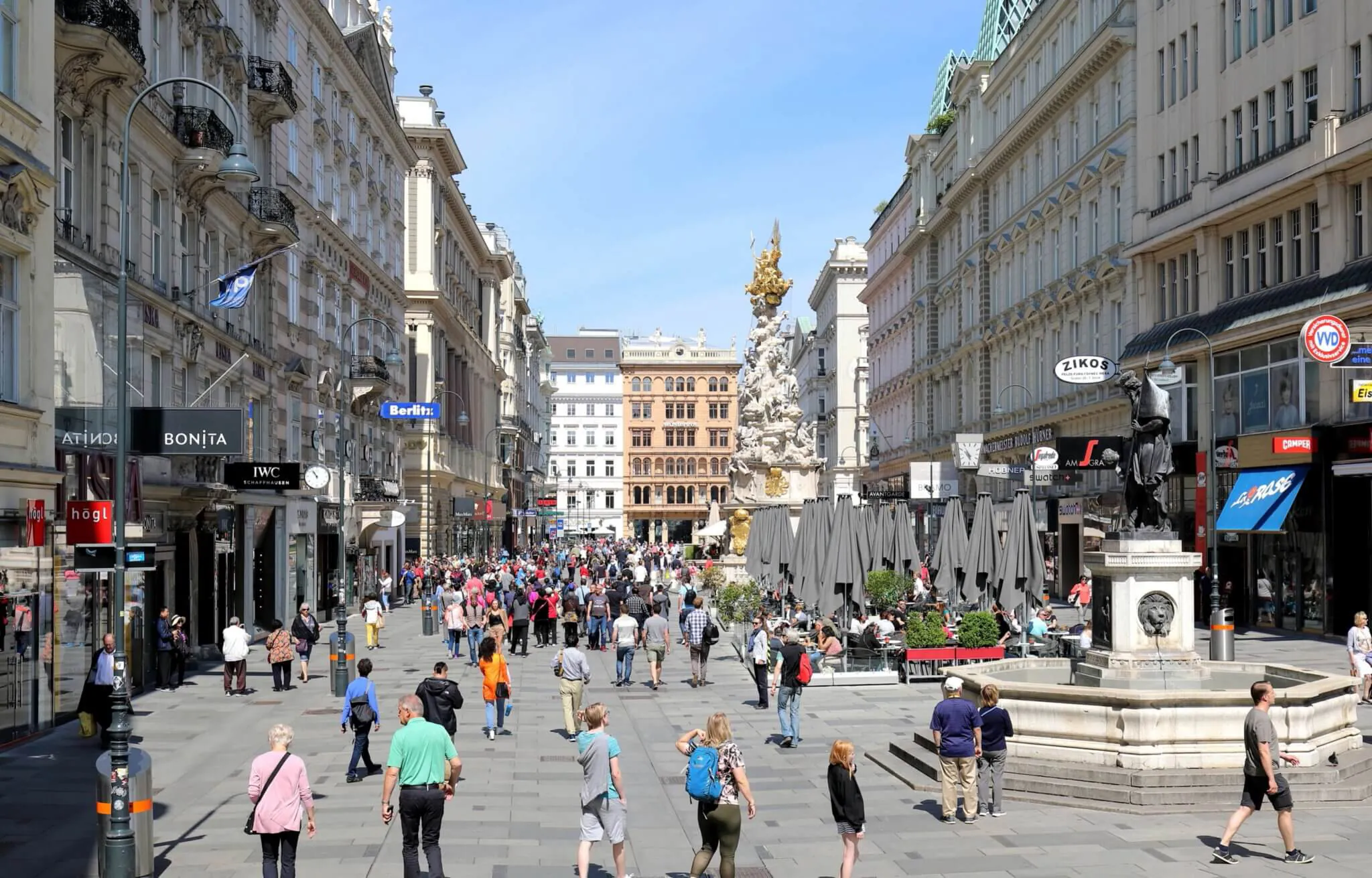Una vivace area pedonale di una città europea offre negozi, caffè e architettura storica. Le persone passeggiano, fanno acquisti e cenano all'aperto. Una colonna imponente e varie statue sono visibili in mezzo alla frenetica scena urbana.  