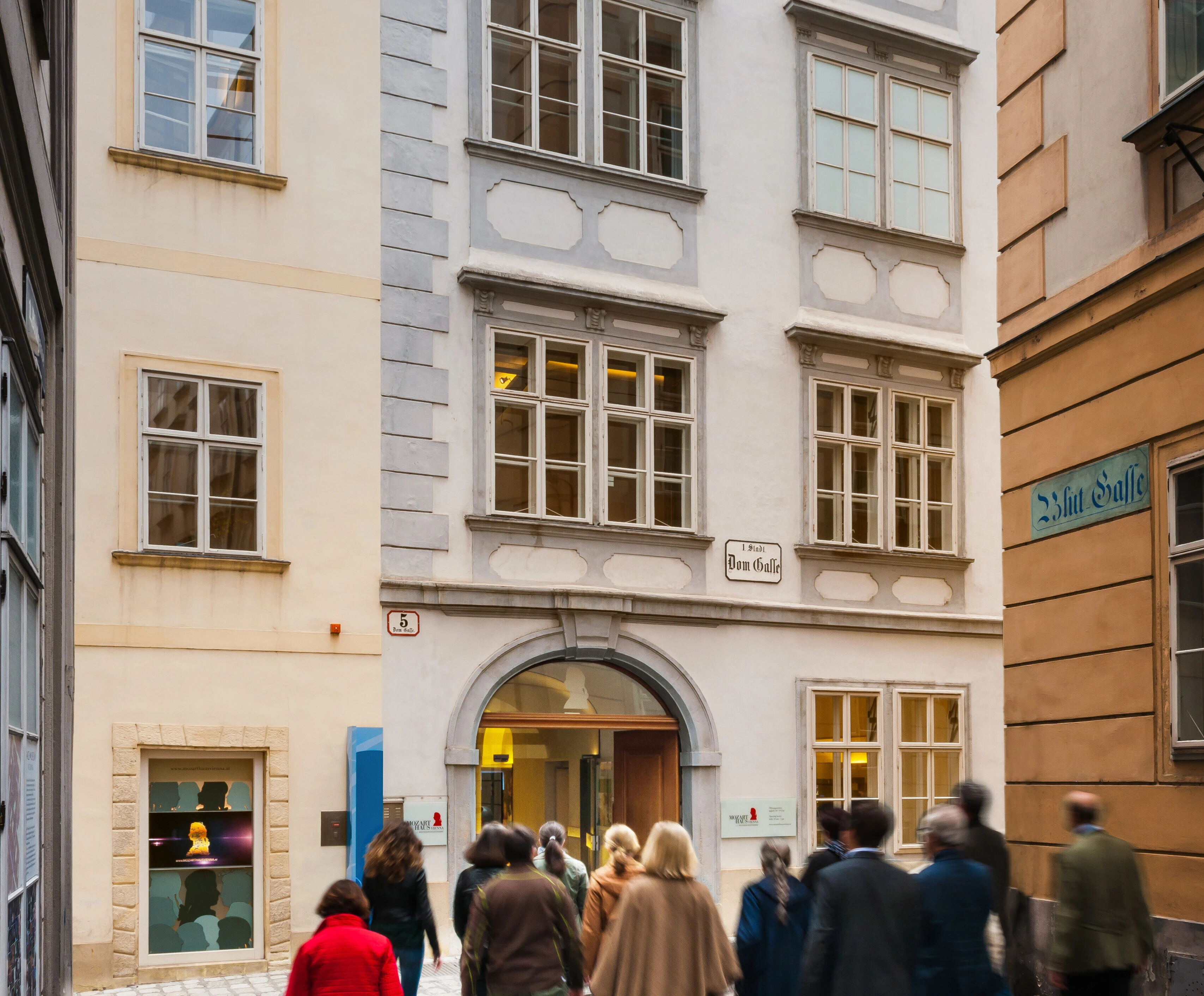 Un gruppo di persone cammina lungo una strada stretta fiancheggiata da edifici a più piani con finestre decorate. Un'insegna è appesa sopra l'ingresso di uno degli edifici. È giorno e la strada è piena di attività.  