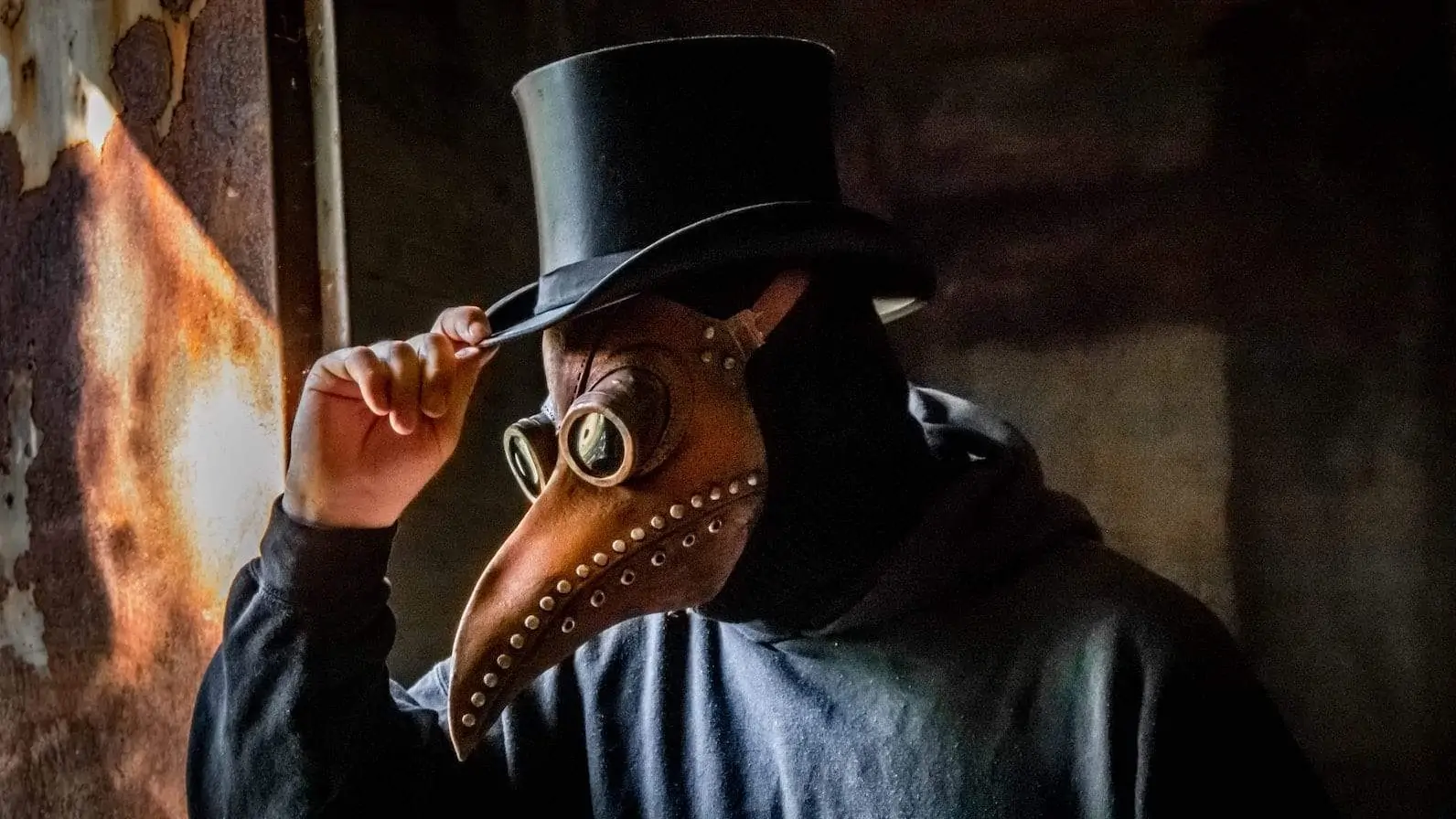 Eine Person, die eine vogelförmige Pestarztmaske mit langem Schnabel und Schutzbrille trägt, rückt einen hohen schwarzen Zylinder zurecht. Die Maske ist mit Metallnieten verziert und der Hintergrund scheint schwach beleuchtet, was auf eine mysteriöse oder historische Kulisse schließen lässt.