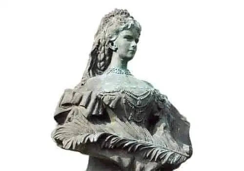 Statue einer Frau in aufwendiger Kleidung aus dem 19. Jahrhundert, mit aufwendigem Schmuck und einem Rüschenkleid mit blattähnlichen Details. Ihr Haar ist mit einem kunstvollen Kopfschmuck geschmückt. Die Skulptur steht vor einem schlichten Hintergrund.