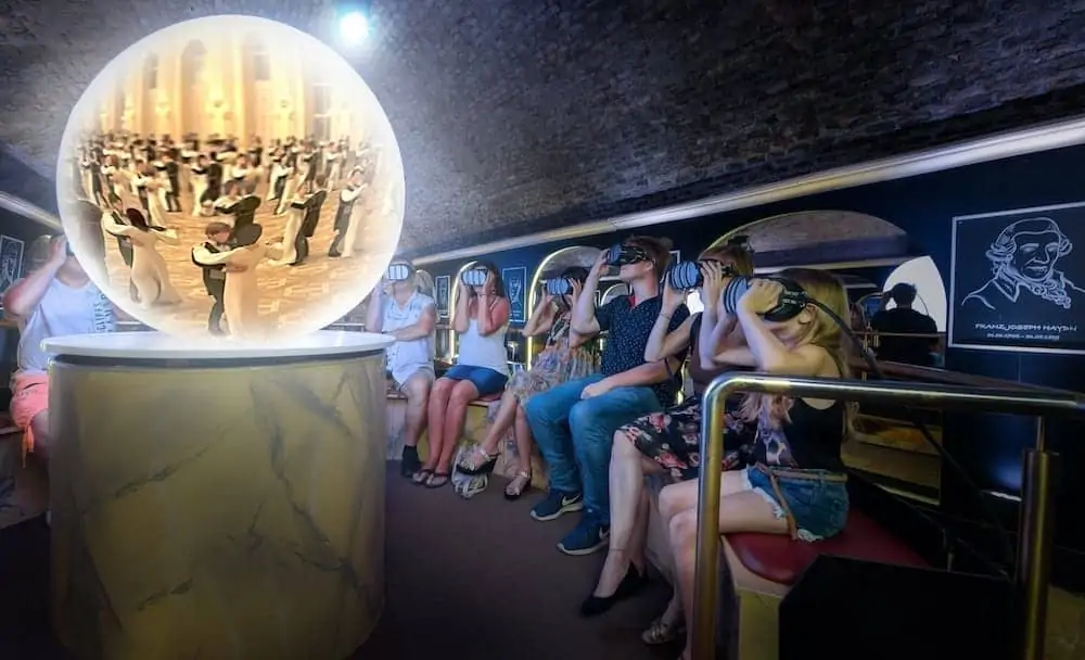 Des personnes sont assises dans une pièce et utilisent des casques de réalité virtuelle. Une grande sphère lumineuse devant eux montre la scène d'un couple dansant, entouré de spectateurs en tenue de soirée. Le décor est moderne et doucement éclairé.