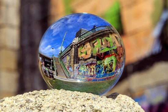 Eine durchsichtige Glaskugel ruht auf einem Felsen und spiegelt eine mit bunten Graffiti bedeckte Wand, eine Treppe und den Himmel wider, wodurch eine verzerrte, künstlerische Ansicht einer städtischen Szene entsteht.