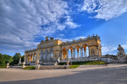 In einem offenen, sonnendurchfluteten Garten steht ein großer, gelber Pavillon im Barockstil mit gewölbten Säulen und Statuen vor einem teilweise bewölkten blauen Himmel.