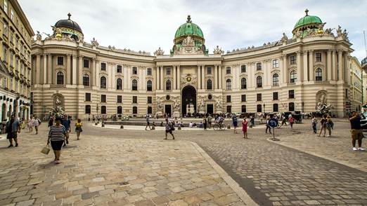 People walk across a cobblestone square in front of the magnificent, historic Hofburg Palace in Vienna, Austria, with its ornate architecture and green domes under a partly cloudy sky.