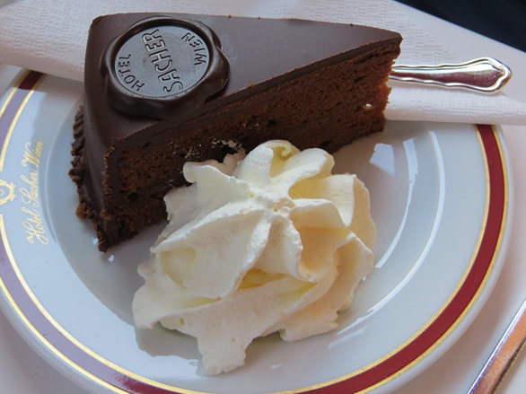 A slice of chocolate cake with a chocolate seal, served on a white plate with a swirl of whipped cream and a fork resting on a napkin.