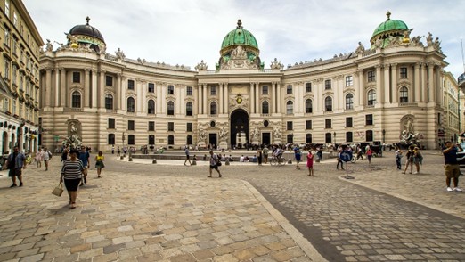 People walk across a cobblestone square in front of the Hofburg Palace in Vienna, Austria. The historic baroque building has a large curved façade and green domed roofs under a cloudy sky.