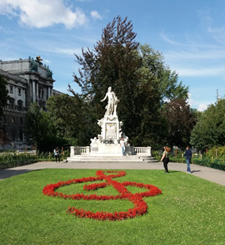 Eine weiße Marmorstatue steht auf einem Sockel in einem Park. Im Vordergrund befindet sich ein rotes Blumenbeet in Form eines Violinschlüssels, in dessen Nähe mehrere Personen spazieren gehen.