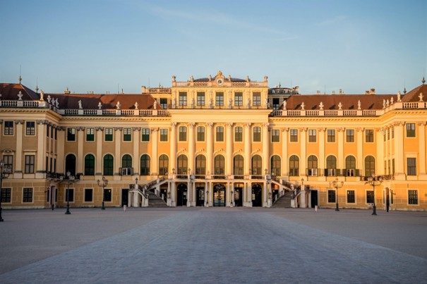 Ein prächtiger gelber Barockpalast mit zahlreichen Bogenfenstern, dekorativen Säulen und einem zentralen Eingang vor einem klaren blauen Himmel, dessen Fassade von sanftem Sonnenlicht erhellt wird.