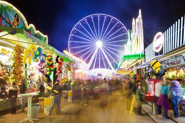 Ein hell erleuchteter nächtlicher Jahrmarkt mit einem leuchtenden Riesenrad im Hintergrund, bunten Ständen, ausgestopften Tierpreisen und Menschenmassen, die die lebhafte Mittelstraße entlanglaufen.