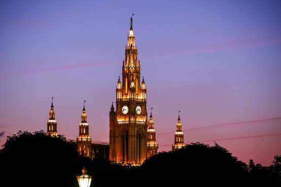 Beleuchteter gotischer Turm und Turmspitzen des Wiener Rathauses vor einem violett-rosa Abendhimmel, teilweise eingerahmt von dunklen Baumsilhouetten.