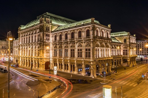 Night view of the illuminated Vienna State Opera with its ornate architecture and arched windows, with the light trails of cars visible on the surrounding streets.