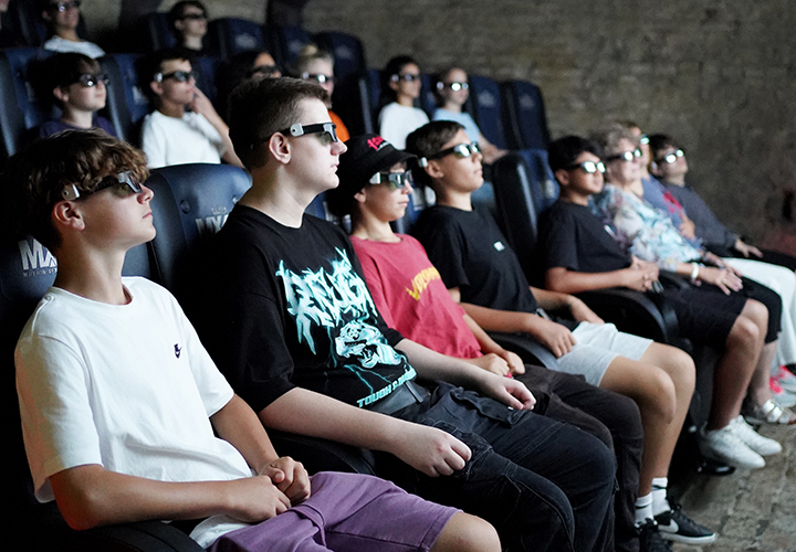 Eine Gruppe junger Leute mit 3D-Brillen sitzt in Reihen von Kinositzen und sieht sich aufmerksam einen Film auf einer großen Leinwand in einem schwach beleuchteten Kino an.