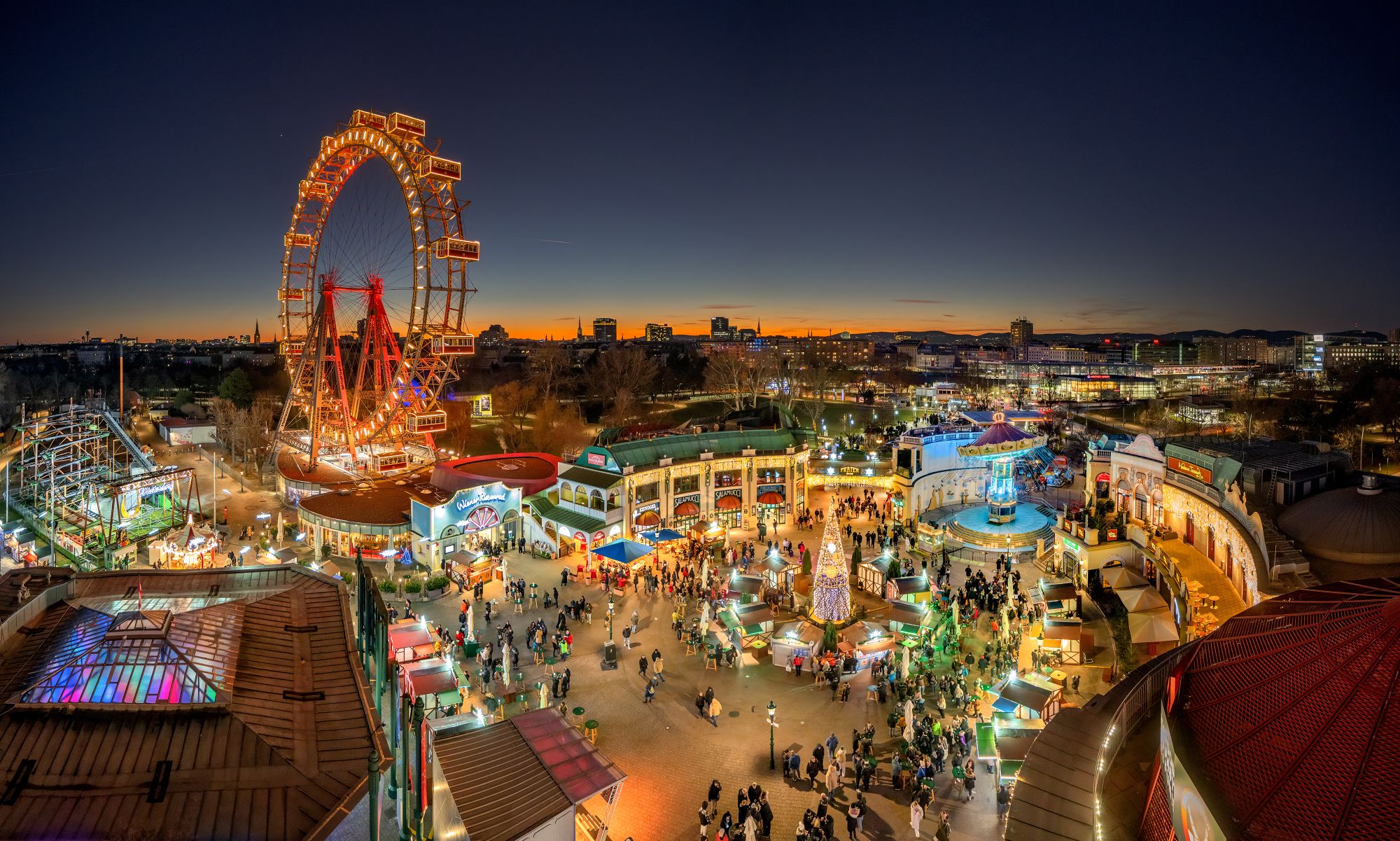 Ein hell erleuchteter Wintermarkt-Vergnügungspark in der Abenddämmerung mit einem großen Riesenrad, bunten Attraktionen und Menschenmassen, die die festliche Atmosphäre unter dem sich verdunkelnden Himmel mit einem glühenden Sonnenuntergang am Horizont genießen.