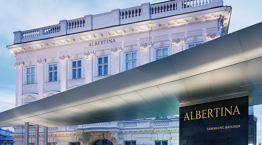 Великолепное историческое белое здание с надписью ALBERTINA, украшенное витиеватыми деталями. На переднем плане - современное здание и черная вывеска, на которой также написано ALBERTINA. Небо чистое и светло-голубое.
