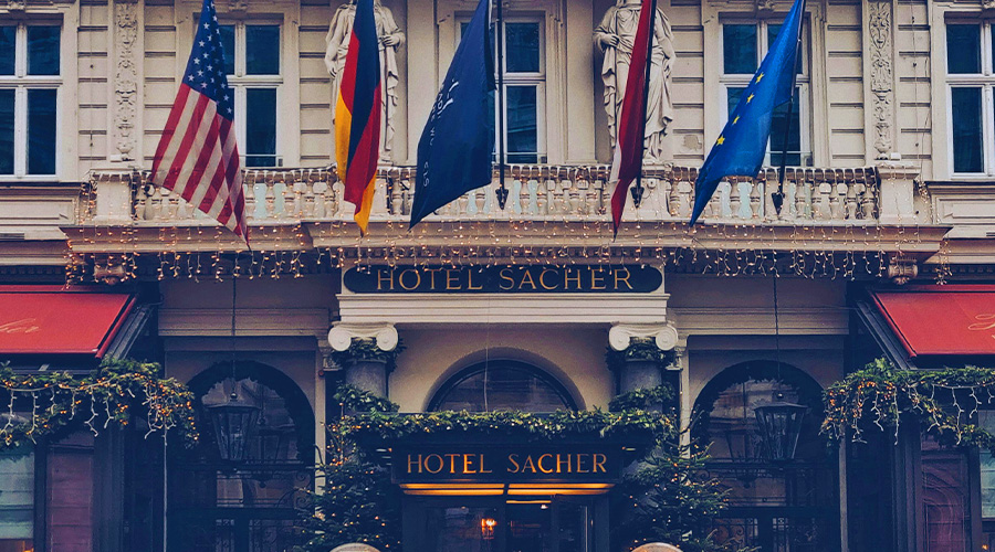 Вход в отель Sacher, украшенный гирляндами и сказочными огнями. Над входом вывешено несколько флагов, в том числе флаги США, Германии, Австрии и Европейского Союза.