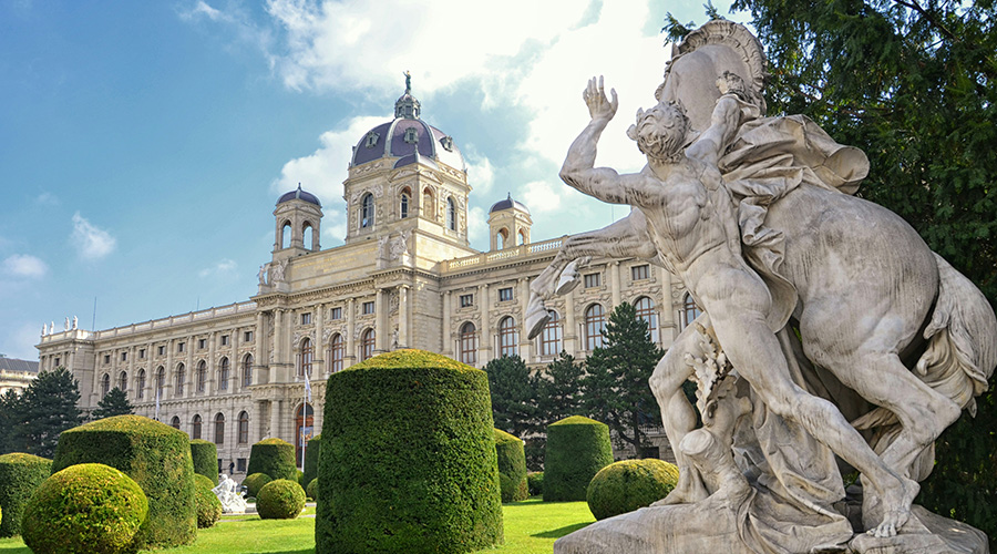 Большое историческое здание с центральным куполом и арочными окнами стоит за ухоженными садами и драматической мраморной статуей двух фигур под ярко-голубым небом.