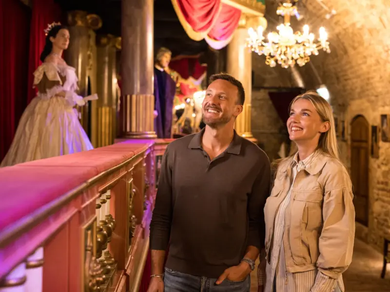 Ein Mann und eine Frau blicken voller Bewunderung zu einem reich verzierten Theaterbalkon mit kostümierten Figuren hinauf.