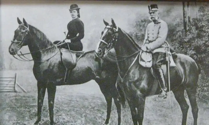 Ein altes Schwarzweißfoto zeigt eine Frau und einen Mann in formeller Kleidung, die nebeneinander im Freien reiten, mit Bäumen im Hintergrund. Die Frau sitzt im Damensattel, der Mann trägt Uniform und Hut.