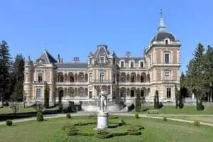 Velký, zdobený palác s detailně propracovaným kamenickým zdivem, klenutými okny a centrální kopulí, obklopený udržovanými trávníky, topiary a sochou v popředí, na pozadí jasně modré oblohy.
