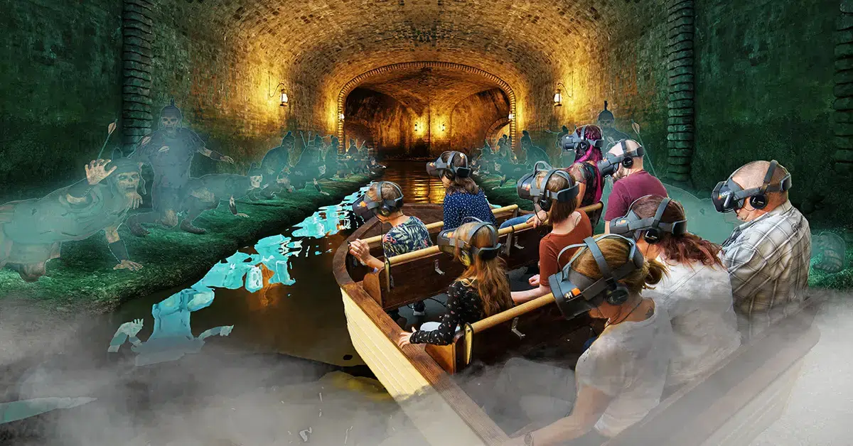 Eine Gruppe von Menschen mit VR-Headsets sitzt in einem Boot, das durch einen dunklen, nebligen Tunnel treibt, umgeben von geisterhaften, leuchtenden Gestalten und einer unheimlichen unterirdischen Landschaft.