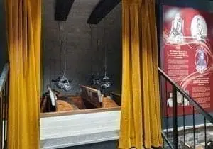 A small room with golden curtains drawn to one side reveals two vintage-style wooden electric chairs under hanging metal head covers. On the wall to the right is an information board with text and pictures.