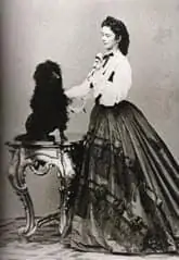 Eine Frau in einem langen, kunstvollen viktorianischen Kleid steht neben einem kleinen Tisch und hält die Pfote eines flauschigen Hundes, der auf dem Tisch sitzt. Das Foto ist schwarz-weiß.