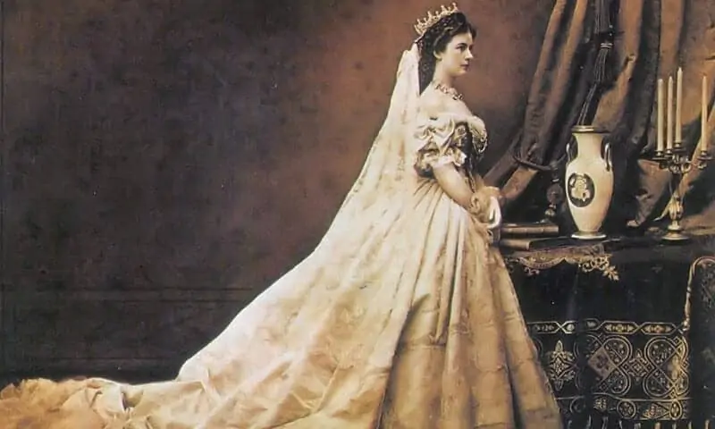 Eine Frau in einem kunstvollen Kleid und mit Krone aus dem 19. Jahrhundert steht im Profil neben einem dekorierten Tisch mit einer Vase und einem Kandelaber, ihr Schleier und ihr Kleid wehen hinter ihr her.