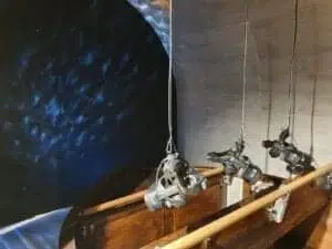 Three theater spotlights hang on cables above the wooden seats and are directed at a large, circular, blue-black structured background on the left. The scene is reminiscent of a stage or an auditorium.